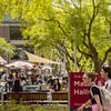 Students walking by a way finding sign in a tree-lined outdoor dining plaza with tables and umbrellas.